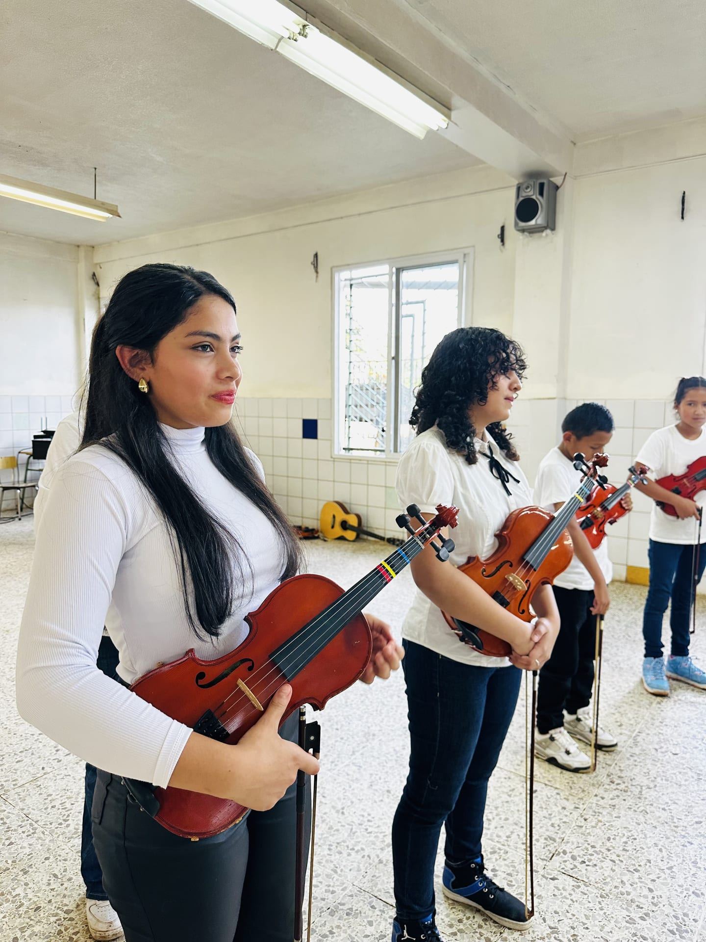 Dos mujeres jóvenes sostienen sus violines en un aula, con niñas y niños de pie detrás de ellas