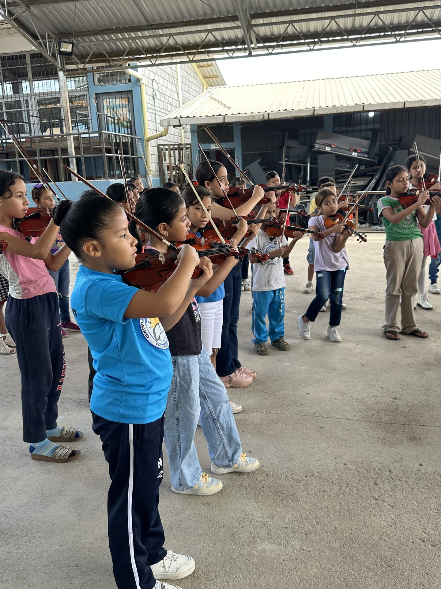 Una fila de niñas y niños toca el violín al aire libre, en un ensayo en el patio del centro educativo
