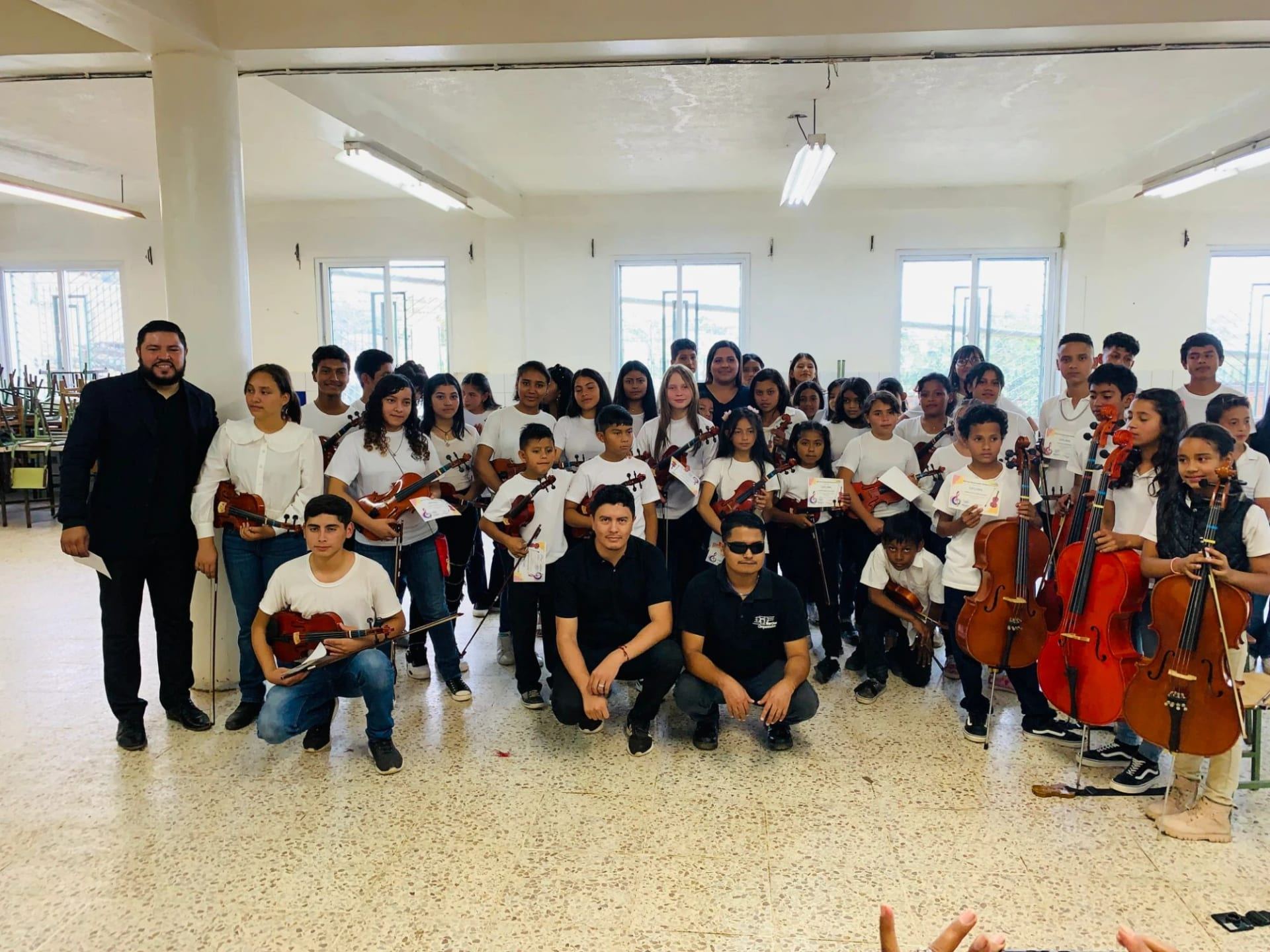 Docentes y alumnos del proyecto posan juntos en un aula con sus violines y violonchelos; varios alumnos sostienen diplomas