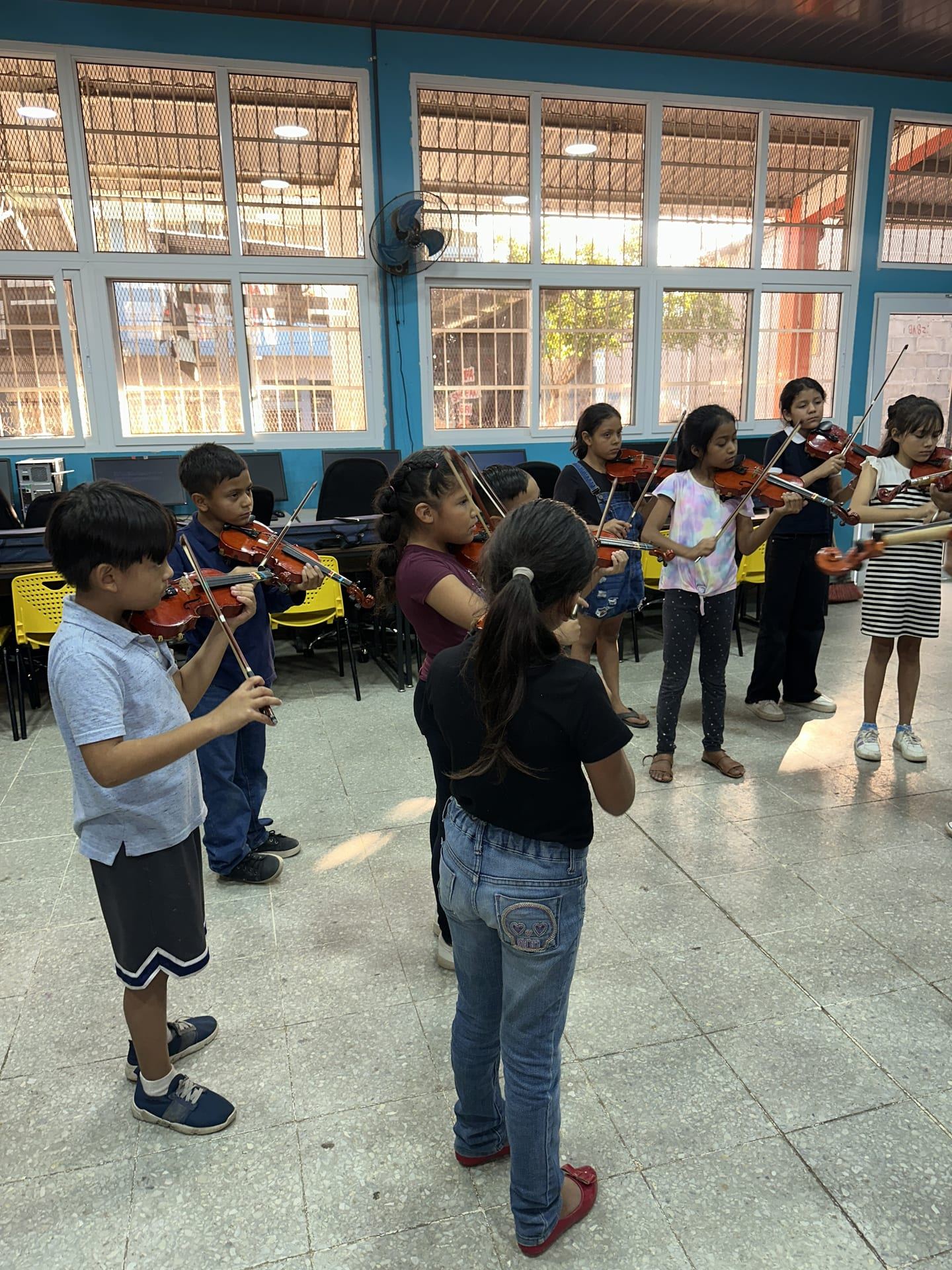 Niñas y niños tocan el violín en un aula con grandes ventanales
