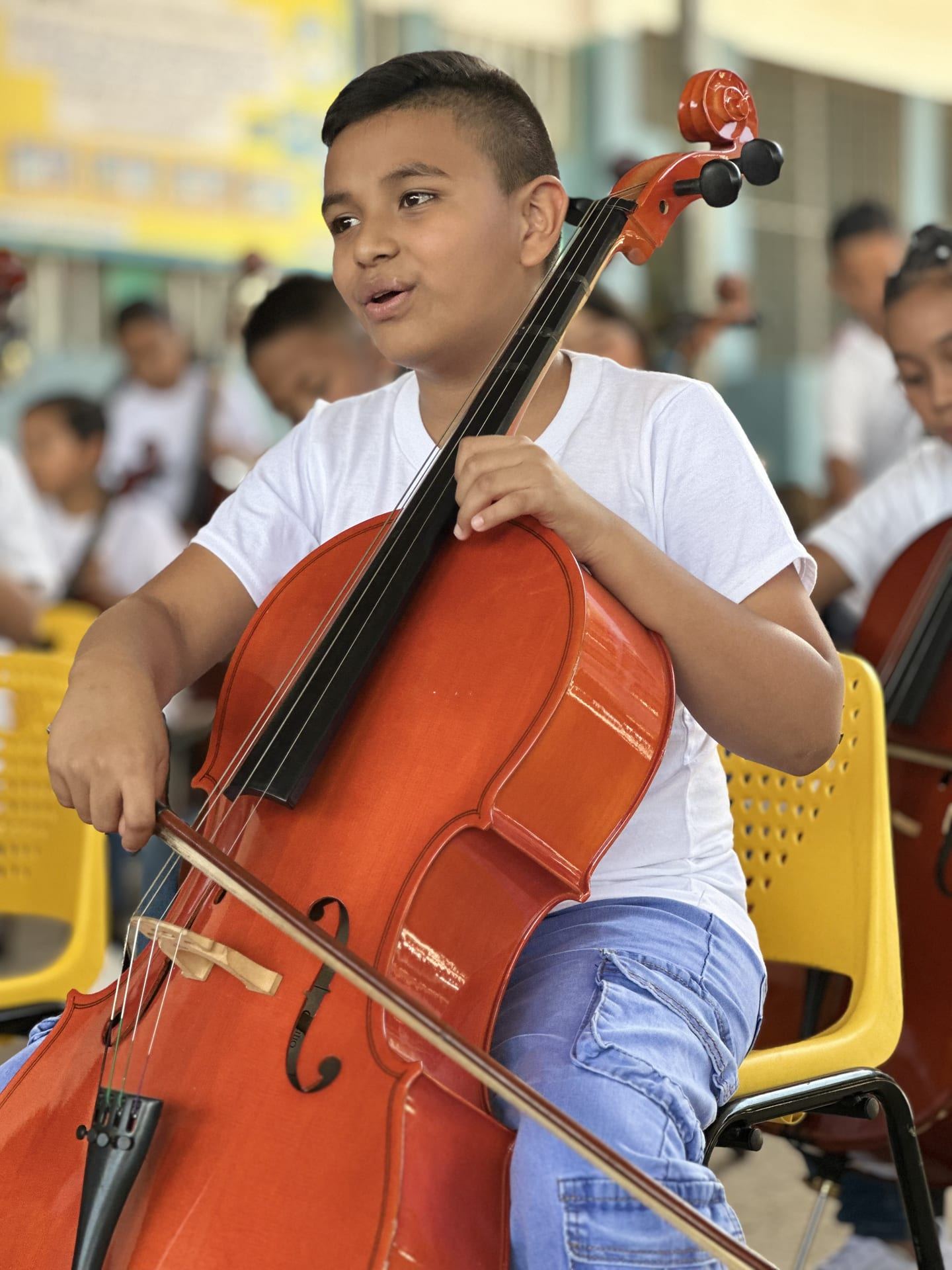 Un niño toca el violonchelo durante un concierto en la sede del proyecto, con otros alumnos desenfocados al fondo