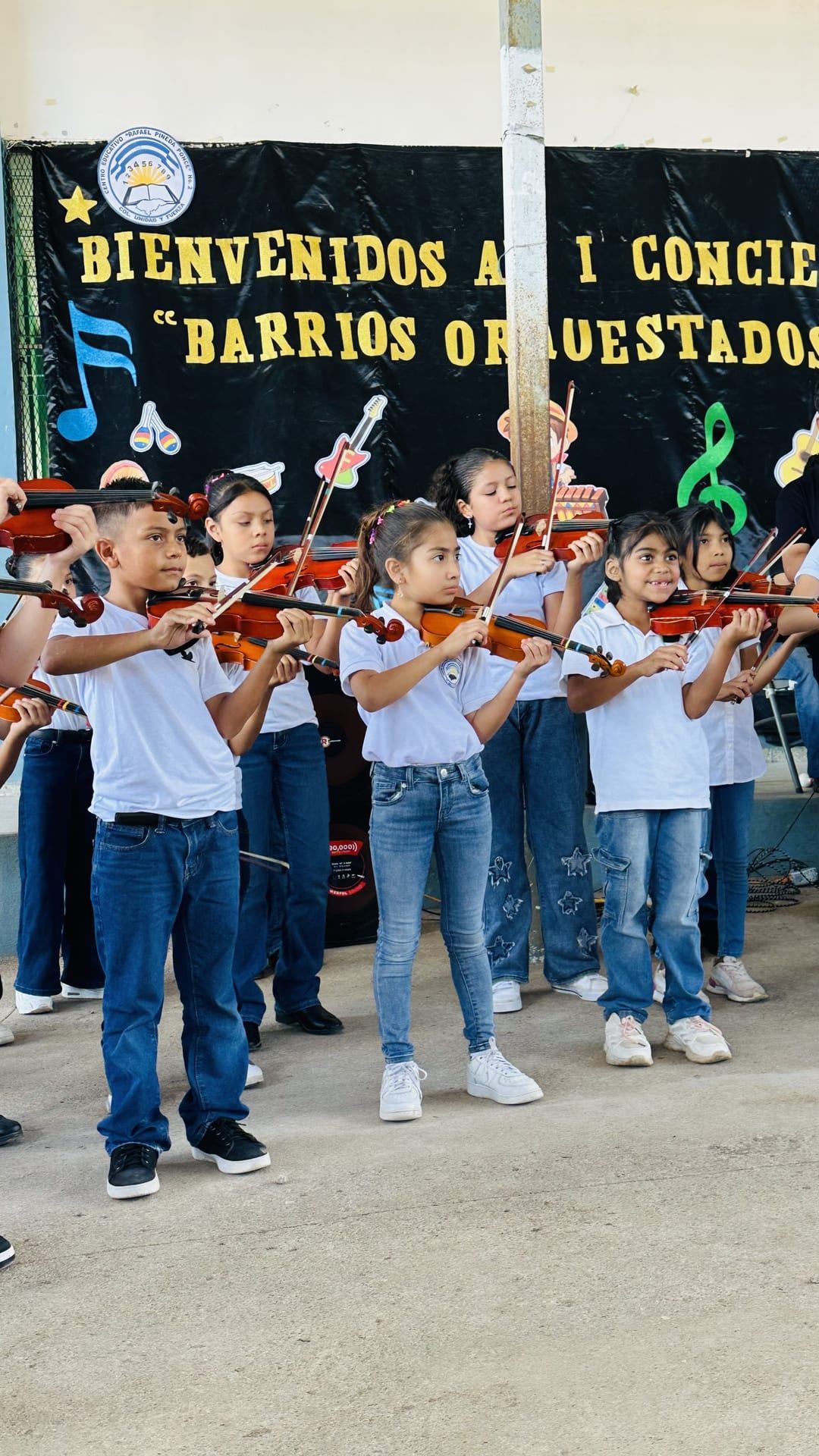Un grupo de niñas y niños toca el violín frente al cartel «Bienvenidos al I Concierto Barrios Orquestados»