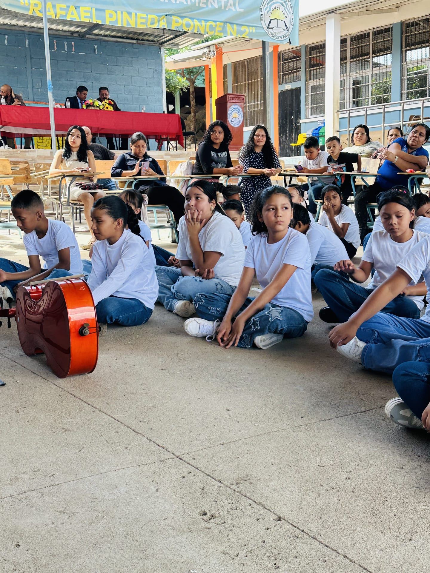Niñas, niños y adultos sentados como público del concierto; en primer plano, un violonchelo apoyado