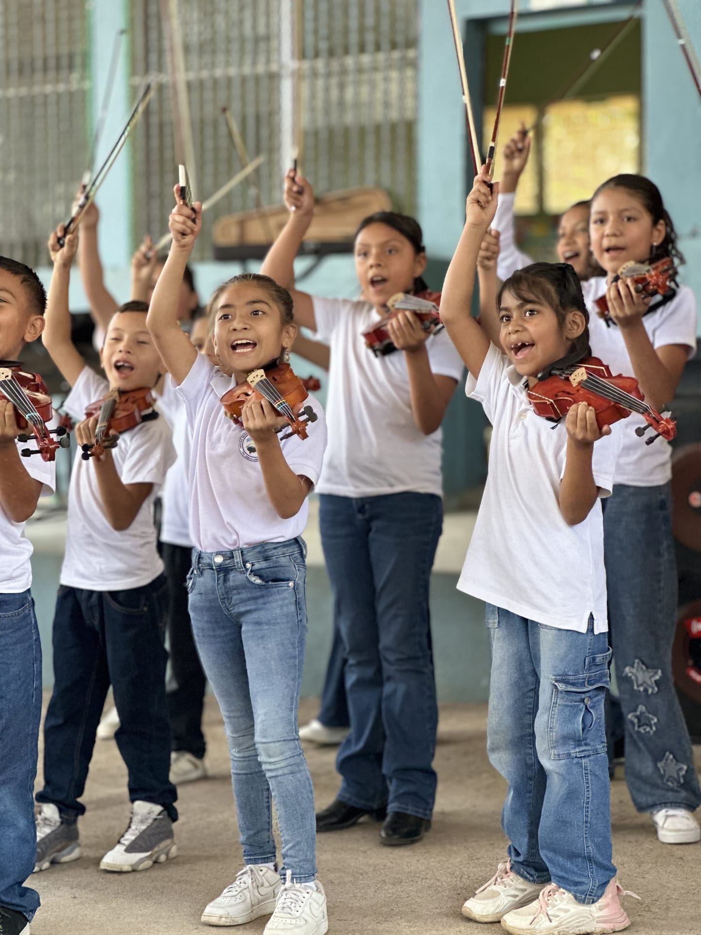 Niñas y niños con camisetas blancas levantan sus violines y arcos, cantando y sonriendo